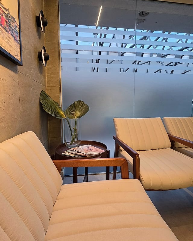 Sala de espera da clínica, com poltronas claras de madeira, mesa de apoio e uma parede envidraçada
